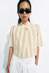Blazer cropped de manga corta de Zara Woman Collection, confeccionado en hilatura con algodón, con diseño a rayas verticales en tonos beige y blanco. Cuenta con cuello subido, bolsillos de vivo en el delantero, forro interior y cierre frontal con botón.