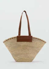 Bolso tipo cesto de tamaño grande, confeccionado en tejido de fibra natural con diseño ensanchado hacia arriba, asas largas y detalles de cuero marrón en el borde superior y la base de las asas.