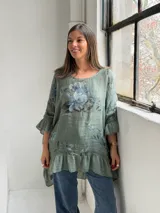 Blusa verde de lino con estampado floral en tonos azules y grises, escote redondo, mangas 3/4 con volados y ruedo asimétrico con volado fruncido.