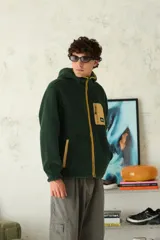 Hoodie de polar color verde oscuro con cierre frontal completo y detalles en color mostaza. Presenta un bolsillo superior con cierre y bolsillos laterales, capucha integrada y diseño de corte relajado.