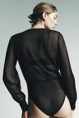 Body negro semitransparente con escote pico profundo, manga larga abullonada con puños ajustados y cierre a presión en la entrepierna. Presenta detalles de volantes y ribetes de puntilla en contraste en el escote y el borde inferior.