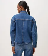Campera de jean azul de corte recto y holgado, con cuello camisero, cierre frontal de botones metálicos dorados, bolsillos frontales con solapa y mangas largas con puños abotonados.