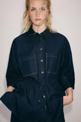 Camisa de denim azul oscuro con cuello solapa, mangas amplias por debajo del codo y bolsillos de parche en el frente. Cuenta con cintura ajustable mediante cordón y cierre frontal con botones metálicos.