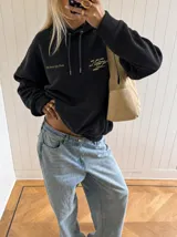 Canguro negro de corte oversize con capucha y bolsillo frontal. Presenta un diseño gráfico circular estampado en color beige en la espalda con tipografía estilizada y detalles de texto en la parte inferior.