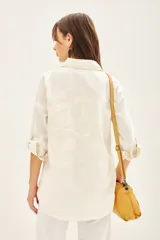 Campera de gabardina color crudo con cuello camisero, mangas largas regulables con tira y botón, y cierre frontal con botones ocultos. Presenta parches bordados decorativos en el delantero y la espalda, además de bolsillos laterales tipo ojal.