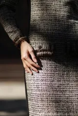 Vestido midi de tweed en tonos beige y marrón, con cuello redondo, manga larga y detalles de flecos en el cuello, puños y bolsillos frontales. Presenta paneles laterales en color negro que aportan estructura y un ajuste entallado.