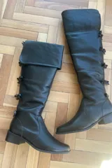 Botas bucaneras de cuero vacuno color negro, con diseño de caña alta ajustable mediante solapa superior y detalle de lazos laterales.