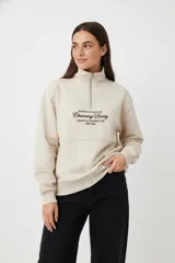 Buzo de algodón color beige claro (hielo) con cuello alto y cierre parcial frontal. Presenta un diseño holgado con franjas horizontales a la altura del pecho y texto estampado en color marrón oscuro.