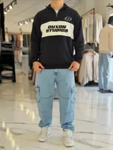 Hoodie de corte clásico con medio cierre y cuello alto, en color marrón con una franja horizontal blanca en el pecho que incluye el logo estampado en negro. Presenta además un pequeño bordado en el hombro izquierdo y puños y cintura elastizados de rib.