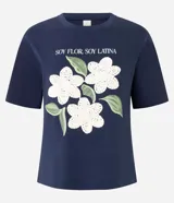 Remera de algodón color blanco, con cuello redondo y manga corta. Presenta un estampado con la frase 'Soy flor. Soy latina' y flores bordadas en relieve con textura tejida.