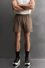 Pantalón corto deportivo color marrón oliva, confeccionado en tejido técnico ligero y elástico, con forro interior. Presenta cintura elástica ajustable con cordón, bolsillos frontales, bolsillo de seguridad trasero con cierre y aberturas laterales en el bajo.