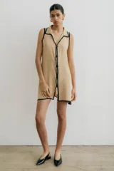 Vestido corto sin mangas color beige, con cuello camisa y cierre frontal con botones negros. Presenta ribetes negros en el cuello, sisas y aberturas laterales.