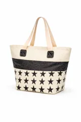Bolso tipo tote de material textil con textura rústica, color beige con estampado de estrellas negras y una franja horizontal de glitter negro. Cuenta con asas largas de color claro y detalles de parches cuadrados negros en la unión de las asas.