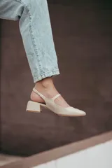 Zapato estilo Mary Jane con diseño destalonado, confeccionado en cuero color beige. Presenta una tira fina con hebilla metálica sobre el empeine, punta redondeada y taco cuadrado forrado de 6 cm de altura.