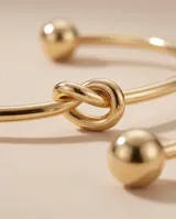 Pulsera tipo esclava rígida de acero con baño de oro, con un diseño minimalista que presenta un detalle de nudo central.