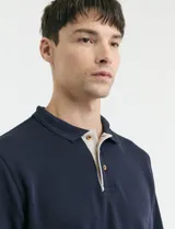 Remera de manga larga con cuello polo, confeccionada en tejido de punto canalé francés. Presenta abotonadura frontal con botones de madera y detalles a contratono en la cartera y el interior del cuello.