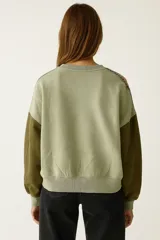 Buzo de tejido de punto color camel, con cuello redondo y mangas largas. Las mangas son de color verde oscuro y presentan una cinta decorativa con patrón étnico en tonos rosados y verdes.