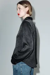 Camisa de satén negro con diseño jacquard de flores tono sobre tono. Presenta escote en pico con solapas, cierre frontal con botones y un broche metálico decorativo en el centro. Tiene mangas largas con puños abotonados.