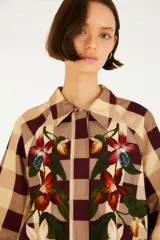 Chaqueta de cuello camisero, manga larga y cierre frontal oculto. Presenta un estampado de cuadros en tonos marrón y beige, complementado con un diseño bordado de flores y hojas en la parte delantera.