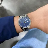 Reloj Casio de acero inoxidable con caja redonda de 38 mm y malla plateada. Presenta una esfera azul con acabado texturizado, índices y manecillas plateadas, y ventana de fecha a las 3.