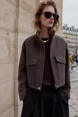 Cazadora de corte corto y diseño estructurado, confeccionada en tejido de paño color marrón. Presenta cuello subido, manga larga, bolsillos delanteros de plastrón con solapa y cierre frontal con cremallera oculta y botones a presión.