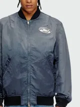 Campera bomber Diesel modelo J-Kepes, color gris azulado con efecto desgastado. Presenta cuello, puños y ruedo en tejido acanalado negro, cierre frontal y un distintivo logo metálico ovalado en el pecho.