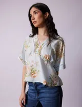 Blusa blanca estampada con flores azules, escote en V y mangas cortas amplias. Detalles de puntilla en escote y mangas.