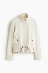Campera corta de lino color marfil con cuello alto, solapa con botones, bolsillos diagonales con solapa, mangas raglán y cordón ajustable en la cintura.