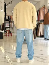 Sweater de punto liviano color beige, con textura marcada, cuello redondo y mangas largas de corte relajado, con pequeño estampado tipográfico negro en el lateral inferior.