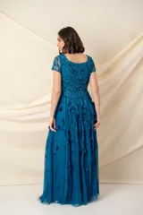 Vestido largo de tul color petróleo, con escote redondo y mangas cortas. Presenta un cuerpo bordado con lentejuelas al tono y una falda con movimiento decorada con apliques de hojas de tela.