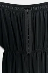 Top negro de manga larga con escote recto estilo off-shoulder, cierre frontal con ganchos metálicos y bajo asimétrico. Las mangas presentan frunces y terminan en puños acampanados con detalle de encaje.