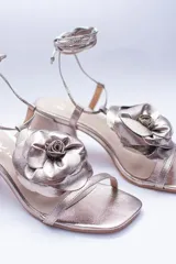 Sandalias de cuero color cobre con taco cuadrado y tira fina sobre los dedos. Presentan una flor grande en el empeine y tiras finas para atar al tobillo.