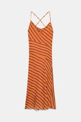 Vestido midi de color naranja con rayas diagonales marrones, escote fluido y tirantes finos cruzados en la espalda con abalorios. Presenta corte evasé con abertura frontal y forro interior.