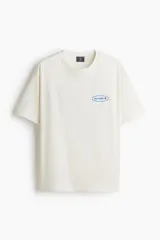 Remera de punto de algodón de peso medio, color blanco hueso, con cuello redondo ribeteado, hombros caídos y bajo recto. Presenta un estampado frontal pequeño con la palabra "uncover" en azul dentro de un óvalo y un símbolo de sol. Tiene un corte holgado.