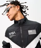 Campera deportiva de poliéster con diseño de bloques de color en negro y blanco. Presenta cuello alto, cierre frontal con cremallera y parches de la NASA y la bandera de Estados Unidos en el pecho.