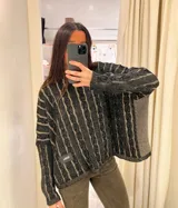 Sweater crop de punto con diseño de trenzas verticales en color beige y camel. Presenta cuello redondo, mangas largas con caída holgada y un parche de cuero sintético con el logo de la marca en la parte inferior.