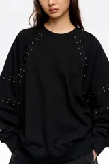Sudadera negra de corte oversize, confeccionada en algodón, con escote redondo y mangas largas tipo ranglán. Presenta detalles de remaches metálicos distribuidos a lo largo de las costuras de las mangas.