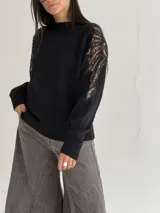 Sweater negro de tejido de punto con cuello redondo y mangas largas. Presenta detalles de transparencias y apliques bordados con pedrería en la parte superior de las mangas.