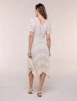 Vestido midi blanco de estilo romántico confeccionado en poliéster con elastano, con detalles de encaje floral. Cuenta con escote cruzado en V, mangas cortas acampanadas, cintura elastizada y falda evasé con terminación asimétrica en picos.