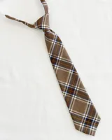 Corbata infantil beige con estampado cuadrillé en tonos marrón, blanco y azul. Tiene un elástico ajustable para sujetar al cuello.