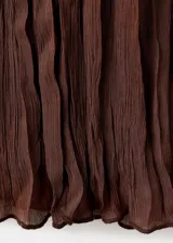 Blusa de color marrón chocolate, confeccionada en tejido plisado con textura. Presenta un diseño de cuello redondo con detalle de lazo, mangas largas amplias con puños acampanados y un corte holgado con volados sutiles en la parte frontal.