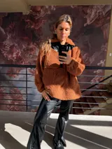 Canguro de peluche color marrón anaranjado con cuello alto y detalles en cuero sintético negro en la parte superior del pecho y cuello. Tiene cierre parcial con botones a presión y dos bolsillos frontales.