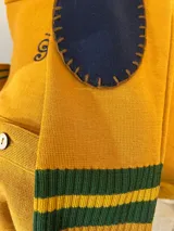 Cárdigan de punto de lana merino color mostaza con cuello tipo polo, cierre frontal de botones y detalles de rayas verdes y amarillas en los puños y el borde inferior. Cuenta con un bordado sutil en el pecho.