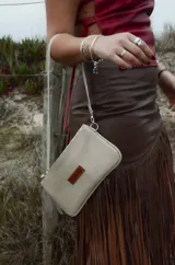 Mini cartera de cuero color beige claro, con asa corta ajustable y cierre superior. Presenta un parche rectangular marrón con la inscripción grabada en la parte frontal.