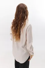 Camisa beige de manga larga con botones marrones y cuello clásico. Presenta un detalle de costura dorada a lo largo de la botonera.