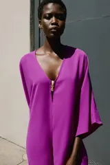 Mono corto de color fucsia, con escote en pico delantero y en la espalda, mangas anchas y largas, y detalle de fornitura dorada en el escote delantero con tiras finas para atar en la espalda.
