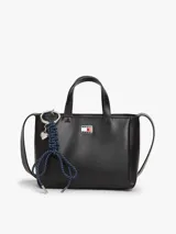 Cartera tote de poliuretano sintético liso color negro de la marca Tommy Jeans, con dos asas superiores y correa ajustable. Presenta cierre de cremallera, compartimento principal e insignia metálica de la marca en la parte delantera. Incluye un adorno colgante extraíble con cordón azul trenzado, dije en forma de corazón y herrajes plateados.