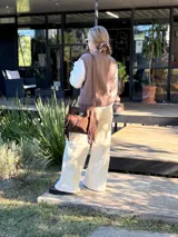 Cartera bandolera de cuero color chocolate, con forro interno de tela y flecos en la parte inferior. Tiene asa larga regulable y cierre superior con botón. Fabricada en Uruguay.