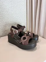 Sandalias de plataforma corrida color marrón, con tiras en el empeine decoradas con perforaciones y tachas metálicas, y tira ajustable con hebilla en el tobillo.