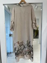 Vestido midi beige de lino, con cierre frontal abotonado, cuello camisero, mangas largas y bolsillos. Presenta un estampado floral en tonos marrones y verdes en la parte inferior, el bolsillo del pecho y el cuello.